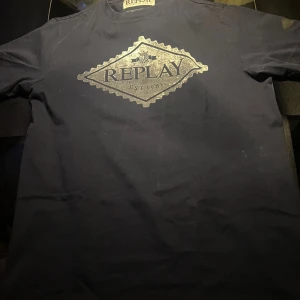 Svart t-shirt från Replay - Snygg svart t-shirt från Replay med ett stort tryck i silver på bröstet. Perfekt för en casual look. Kortärmad och tillverkad i ett mjukt material.
