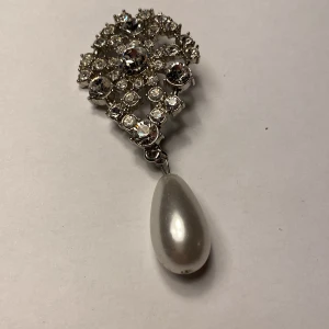Elegant brosch med pärldroppe - Snygg vintage brosch från USA i silverfärg med glittrande stenar och en stor pärldroppe. Perfekt för att ge en touch av elegans till din outfit. Fästes enkelt med en nål på baksidan.