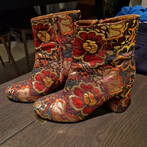 Blommiga boots med klack - Unika boots med blommigt mönster i rött, gult och orange. Skorna har en bekväm klack och dragkedja på sidan för enkel på- och avtagning. Perfekta för att ge en färgglad touch till din outfit.