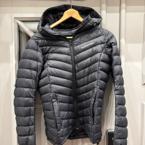 Svart dunjacka från Peak Performance - Snygg och funktionell svart dunjacka från Peak Performance. Jackan har en dragkedja framtill och är tillverkad i Pertex Quantum-material för extra hållbarhet och värme. Perfekt för kyliga dagar med sin långa ärm och stilrena design.