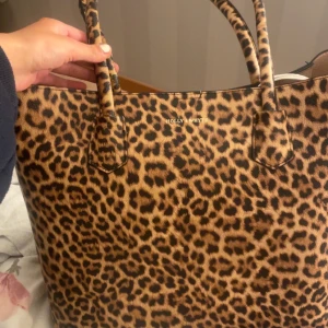 Leopardmönstrad väska från Holly & Whyte - Snygg leopardmönstrad väska från Holly & Whyte. Perfekt för att ge din outfit en djärv touch. Rymlig insida med plats för alla dina nödvändigheter. Stilren design med två handtag för enkel bärning.