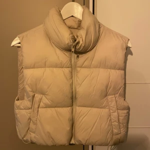 Beige dunväst med dragkedja - Snygg beige dunväst med hög krage och dragkedja framtill. Perfekt för kyligare dagar och ger en stilren look. Västen har en quiltad design och praktiska fickor på sidorna.