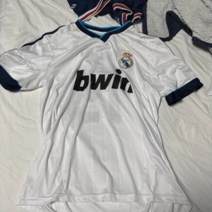 Vit Real Madrid fotbollströja - Säljer en vit Real Madrid fotbollströja med korta ärmar och blå detaljer. Tröjan har 'bwin' som sponsorlogga på framsidan och 'Modrić 19' tryckt på baksidan. Perfekt för fans av laget! Pris kan diskuteras 