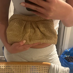 Guldig kuvertväska - Snygg guldig kuvertväska med flätad design. Perfekt för att ge en glittrig touch till din outfit. Väskan har en mjuk insida och är lätt att bära med sig.