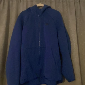 Blå hoodie från Nike - Säljer en blå hoodie från Nike med dragkedja och huva. Perfekt för kyliga dagar med sin bekväma passform och praktiska fickor framtill. En klassisk och stilren design som passar till det mesta.