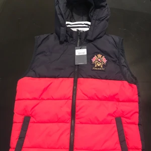Marinblå och röd väst från Hackett - Oanvänd, snygg väst från engelska Hackett i marinblått och rött med broderad logga på bröstet. Västen har en praktisk  avtagbar huva och dragkedja framtill. Den har två sidofickor men utan dragkedja eller knapp.Perfekt för mindre kyliga  dagar på hösten och tidig vår. Storlek 13-14 år men liten i storlek och är snarare 10-12 år. 