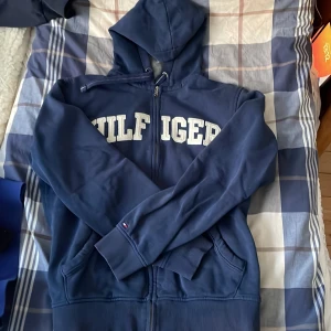 Blå hoodie från Tommy Hilfiger - Säljer en snygg blå hoodie från Tommy Hilfiger med dragkedja och stor logga på bröstet. Perfekt för en avslappnad stil. Den har långa ärmar och en praktisk huva.