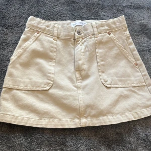 Beige kjol från Zara - Snygg beige kjol från Zara i kort modell. Kjolen har praktiska fickor fram och stängs med knapp och dragkedja. Perfekt för en stilren look.