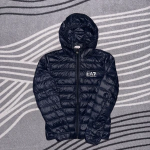Svart pufferjacka från EA7 Emporio Armani - Snygg svart pufferjacka från EA7 Emporio Armani med huva och dragkedja. Perfekt för kyligare dagar med sin quiltade design som håller värmen. Jackan har en stilren look med EA7-logga på bröstet.