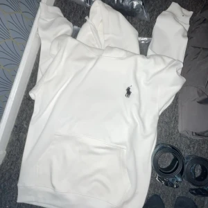 Vit hoodie från Ralph Lauren - Säljer en stilren vit hoodie från Ralph Lauren med det klassiska logotypbroderiet på bröstet. Tröjan har en stor ficka framtill och en bekväm huva. Perfekt för en avslappnad look.