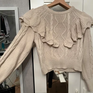 Beige stickad tröja från ONLY - Säljer en beige stickad tröja från ONLY med volangdetaljer på axlarna. Tröjan har ett fint kabelstickat mönster och långa ärmar. Perfekt för en mysig och stilren look.