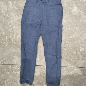 Blå kostymbyxor i linne - Snygga blå linnebyxor med en klassisk passform. Perfekta för en stilren look, oavsett tillfälle. Byxorna har en midwaist design och stängs med knappar. De är tillverkade i ett lätt och luftigt material som passar bra för varmare väder.
