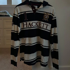 Randig pikétröja från Hackett - Snygg randig pikétröja från Hackett i svart och vit med broderad logga på bröstet. Tröjan har långa ärmar och en klassisk krage med knappar. Perfekt för en stilren look.