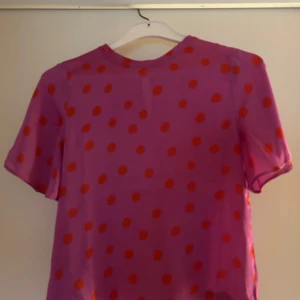 Lila prickig blus - Säljer en lila blus med röda prickar. Blusen har korta ärmar och en rund halsringning. Perfekt för att liva upp garderoben med lite färg och mönster!
