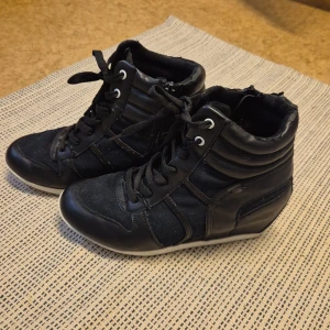 Svarta sneakers med kilklack från VOX - Snygga svarta sneakers från VOX med kilklack och snörning. Skorna har en sportig design med dragkedja på sidan för enkel på- och avtagning. Perfekta för en stilren look.