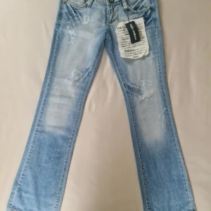 Ljusblå jeans från D&G - Snygga ljusblå jeans från D&G med slitna detaljer och nitar längs midjan. De har en klassisk femficksdesign och en knapp med D&G-logga. Perfekta för en trendig look. Storlek 42 och är nya med tagg