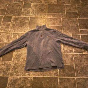 Blå tröja från Ralph Lauren - Säljer en stilren grå tröja från Ralph Lauren med dragkedja och orange broderad logga på bröstet. Tröjan har långa ärmar och en hög krage, perfekt för kyligare dagar.