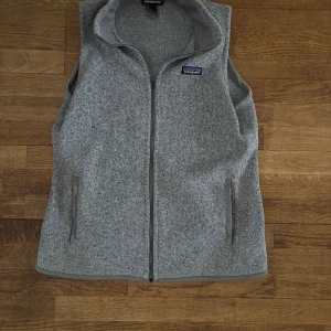 Grå fleeceväst från Patagonia - Säljer en stilren grå fleeceväst från Patagonia. Västen har en mjuk och bekväm passform, perfekt för lager-på-lager under kyligare dagar. Den är i storlek L i dam och de motsvarar M i herr storlek