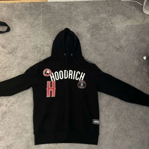 Hoodrich hoodie - Säljer en svart hoodie från Hoodrich med tryck i vitt och rött på framsidan. Den har en justerbar huva med dragsko och en stor magficka. Perfekt för en avslappnad stil.