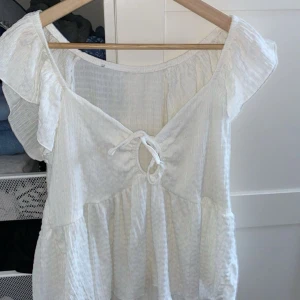 Vit blus med knytning - Charmig vit blus med korta ärmar och knytning framtill. Blusen har en luftig och lätt design med volangdetaljer vid ärmarna, perfekt för varma dagar.