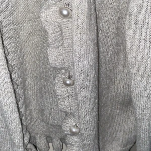 Grå stickad kofta med pärldetaljer - Säljer en elegant grå stickad kofta med volangdetaljer och pärlknappar. Perfekt för att ge en stilren touch till din outfit. Koftan har långa ärmar och en mjuk, bekväm passform.