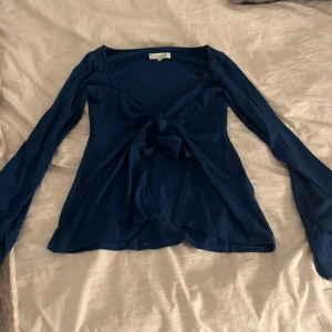 Blå långärmad topp från Design by si - Snygg blå långärmad topp från Design by si med knytning framtill. Perfekt för en stilren look med en touch av elegans. Toppen har en v-ringad design och är tillverkad i ett mjukt material som ger en bekväm passform.