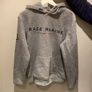 Grå hoodie från Race Marine - Snygg grå hoodie från Race Marine med tryck på bröstet och ärmen. Den har en klassisk känguruficka och en bekväm huva. Perfekt för en avslappnad stil.