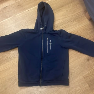 Mörkblå zip hoodie från Sail Racing - Säljer en mörkblå hoodie från Sail Racing med dragkedja och huva. Den har en praktisk bröstficka med dragkedja och märkeslogga på framsidan. Perfekt för kyliga dagar. Ny pris 1000. Skick 8-10 pris kan diskuteras!