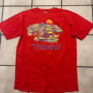 Röd t-shirt med Thailand-motiv - Säljer en röd t-shirt med ett färgglatt Thailand-motiv på framsidan. T-shirten har korta ärmar och en rund halsringning. Perfekt för en avslappnad stil.