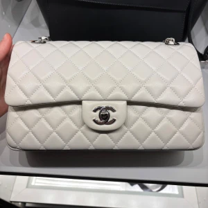 Vit  axelväska  Chanel - Elegant vit axelväska från Chanel i quiltat skinn med silverdetaljer. Väskan har en klassisk design med kedjerem och ikonisk logotyp på framsidan. Insidan har en dragkedja för extra säkerhet och stil.