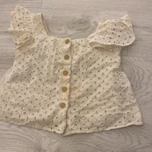 Vit blus från Zara Kids - Söt vit blus från Zara Kids med spetsdetaljer och knappar framtill. Blusen har korta ärmar med volang och ett vackert hålmönster. Perfekt för sommarens varma dagar!