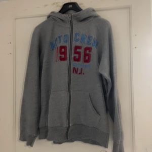 Grå hoodie från H&M - Säljer en grå hoodie från H&M med texten 'AUTO CREW 1956 N.J.' i blått och rött på framsidan. Tröjan har dragkedja och en bekväm huva. Perfekt för en avslappnad stil.
