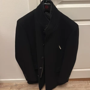 kappa med knappar - Elegant svart kappa med knappar och quiltad insida som går att ta av. Den har en stilren design med en hög krage och en dold dragkedja. Perfekt för kyligare dagar. 