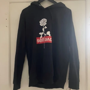 Svart hoodie med rosdetalj - Snygg svart hoodie med en vit ros och texten 'selflove' i rött på framsidan. Perfekt för en avslappnad stil med en touch av attityd. Långärmad och med en klassisk huva.