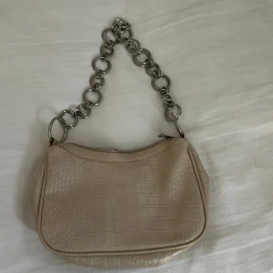 Beige axelväska med kedjedetaljer - Snygg beige axelväska med krokodilpräglat mönster och en unik kedja som axelrem. Väskan har en dragkedja upptill för säker förvaring. Perfekt för att ge din outfit en trendig touch.