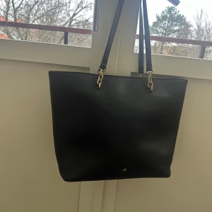 Svart axelväska i skinn - Elegant svart axelväska i skinn med guldfärgade detaljer. Perfekt för en stilren look och rymmer allt du behöver för dagen. Väskan har en enkel design med långa axelremmar för bekväm bärning.