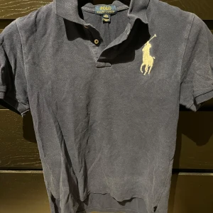 Mörkblå pikétröja från Ralph Lauren - Säljer en klassisk mörkblå pikétröja från Ralph Lauren med korta ärmar och en broderad logga på bröstet. Tröjan har en krage med knappar och en vit siffra på ärmen. Perfekt för en stilren look. Varann är inköpt från Kidsbrandstore. Storleken på varann är barnstorlek XL, 18-20 år. Jag anser att detta motsvarar en storlek S-M i herr storlek. 