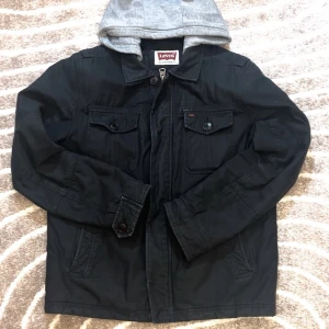 Svart jacka med grå huva från Levi's - Snygg svart jacka från Levi's med grå huva. Jackan har knappar och dragkedja framtill samt två bröstfickor med knappstängning. Perfekt för en casual look.