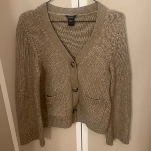 Beige stickad kofta från DKNY - Mysig beige stickad kofta från Lindex med v-ringning och knappar framtill. Koftan har två fickor och långa ärmar. En stilren och bekväm tröja att ha i garderoben.