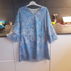 Blommig blå klänning - Charmig blå klänning med ett blommigt mönster och trekvartsärmar. Perfekt för vår och sommar med sin luftiga design och v-ringade hals. Klänningen har en avslappnad passform och volangdetaljer på ärmarna.