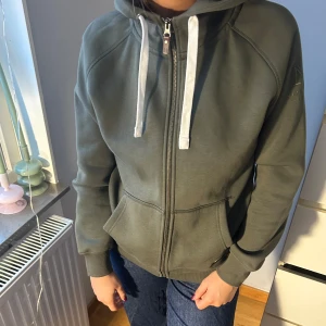 Grön hoodie från Nimbus - Säljer en grön hoodie från Nimbus med dragkedja och justerbar huva. Den har en broderad logga på ärmen och praktiska fickor framtill. Perfekt för en avslappnad stil. NYSKICK