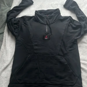 Svart hoodie från Jordan - Säljer en svart hoodie från Jordan med dragkedja och känguruficka. Den har en hög krage och justerbar dragsko vid halsen. Perfekt för en sportig look eller avslappnade dagar. Minimalt hål i armhålan. 