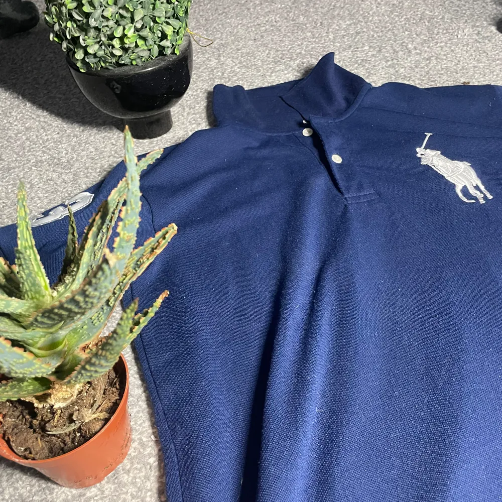 Säljer en snygg mörkblå pikétröja från Ralph Lauren med klassisk krage och knappar. Tröjan har en stor vit logotyp på bröstet och en siffra på ärmen. Perfekt för en stilren look!. Neuletakit & Villapaidat.