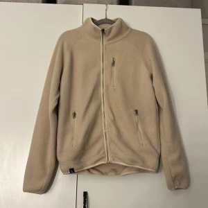 Beige fleecejacka med dragkedja - Mysig beige fleecejacka med dragkedja och hög krage. Jackan har två sidofickor och en bröstficka, alla med dragkedjor. Perfekt för kyliga dagar.