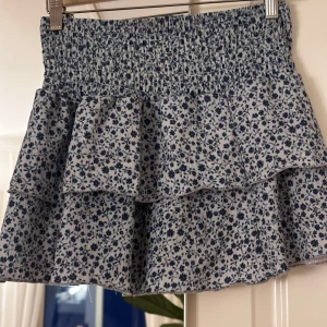 Blommig volangkjol - Söt kortkjol med blått blommönster på vit bakgrund. Kjolen har en elastisk midja och volangdetaljer som ger en lekfull look. Perfekt för varma dagar.