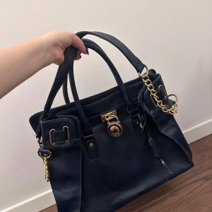 Mörkblå handväska med gulddetaljer Michael kors - Snygg mörkblå handväska med eleganta gulddetaljer och ett lås framtill. Väskan har två handtag och en rymlig insida med mönstrat foder. Perfekt för att bära dina dagliga nödvändigheter med stil.