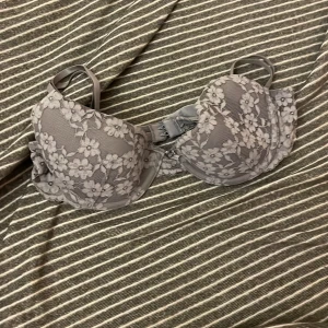 Grå blommig spets-bh - Elegant grå bh med vacker blommig spetsdesign. Den har justerbara axelband och en klassisk hakknäppning bak. Perfekt för att ge en feminin touch till din garderob.
