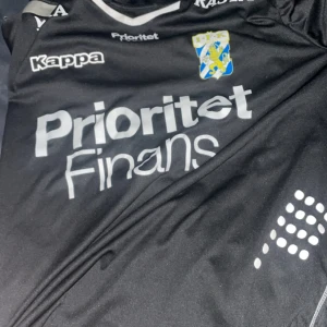 Svart fotbollströja från Kappa - Snygg svart fotbollströja från Kappa med vit text och klubbmärke på bröstet. Perfekt för fotbollsfans som vill visa sitt stöd. Tröjan har korta ärmar och en modern design.