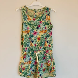 Blommig ärmlös jumpsuit - Snygg och färgglad ärmlös jumpsuit med tropiskt blommönster i grönt, rosa och gult. Perfekt för varma sommardagar med bekväm passform och knytband i midjan.