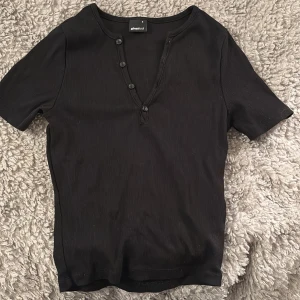 Svart ribbad topp från Gina Tricot - Snygg svart ribbad topp från Gina Tricot med korta ärmar och knappar längs den v-ringade halsen. Perfekt för en stilren look.
